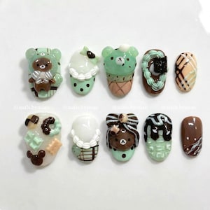 May include: Ten nail art designs with dessert themes, including bear, ice cream, and donut shapes. The designs feature brown, green, and white colours. The nails are displayed on a white surface.