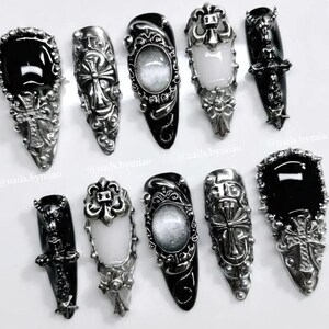 Op de afbeelding: Een assortiment van sierlijke, zilverkleurige nail art ontwerpen. De collectie bevat zwarte en witte ovale stenen, kruisen en ingewikkeld metaalwerk. De nagels zijn gerangschikt op een wit oppervlak, wat een gotische esthetiek laat zien.