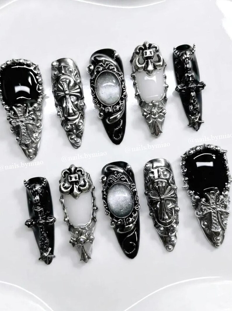 Op de afbeelding: Een assortiment van sierlijke, zilverkleurige nail art ontwerpen. De collectie bevat zwarte en witte ovale stenen, kruisen en ingewikkeld metaalwerk. De nagels zijn gerangschikt op een wit oppervlak, wat een gotische esthetiek laat zien.