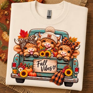 May include: A cream-colored crewneck sweatshirt featuring a graphic of a teal vintage truck carrying three fluffy highland cows. The cows are decorated with a leopard print bow, a knit beanie, and a plaid bow. The truck bed displays the text Fall Vibes.