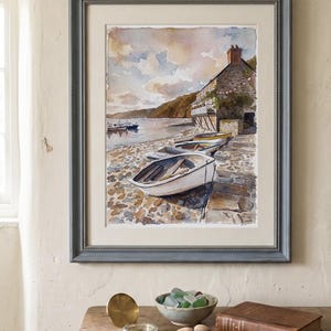May include: A framed watercolor painting depicts a coastal scene with boats on a pebbled shore. The painting is in a gray frame, and the scene includes a building, boats, and a cloudy sky. A wooden table in the foreground holds a compass, bowl, and book.