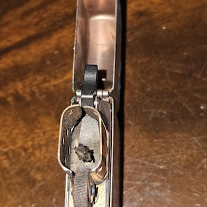 Puede incluir: Un encendedor abierto de estilo vintage con un interior color cobre y un exterior plateado. El mecanismo del encendedor, incluyendo la rueda de pedernal y la mecha, es visible. El encendedor está sobre una superficie de madera.