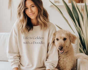 以下が含まれることがあります： 「Life is Golden with a Doodle」の文字が入ったベージュのスウェットシャツ。女性とゴールデン・ドゥードゥル犬が白いソファに座っています。犬は薄茶色で巻き毛です。女性は長く波打つ茶色の髪をしています。