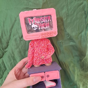 Op de afbeelding: Een roze televisie met de woorden "Monster High" op het scherm is bevestigd aan een roze gebreide trui. De trui ligt op een paarse commode met twee roze lades.