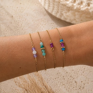 May include: Four gold chain bracelets, each with a unique arrangement of colourful beads. The beads feature shades of purple, turquoise, and blue. The bracelets are shown on a person's arm.