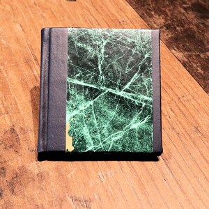 May include: A small, rectangular book with a dark blue spine and cover. The front cover features a marbled pattern in shades of green and white. The book is resting on a wooden surface.