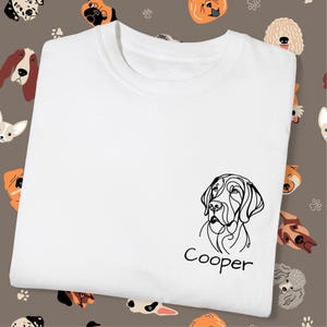 May include: White t-shirt featuring a black line drawing of a dog's face with the name "Cooper" below it. The shirt is folded, and the background has a pattern of various dog faces.