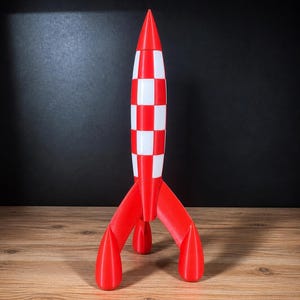 Op de afbeelding: Een rood en wit raketmodel met een geblokt patroon op de romp en drie rode vinnen. De raket heeft een puntige top en wordt tentoongesteld op een houten oppervlak. Het model is een decoratief item.