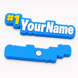 Puede incluir: Un imán personalizado azul, amarillo y blanco con el texto #1 YourName en una fuente gruesa y en relieve. El frente muestra las letras coloridas, mientras que la parte posterior muestra un imán circular de metal plateado en la base.