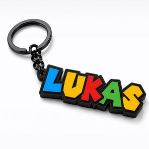 May include: A keychain featuring the name LUKAS in a blocky, multi-colored font with a black border. The letters are blue, yellow, red, green, and yellow, attached to a black metal split ring by a short black chain link connector.