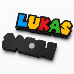 Pu&ograve; includere: Un magnete colorato con il nome "LUKAS" in lettere blu, gialle, rosse e verdi con un contorno nero. Un magnete nero separato con un magnete argentato &egrave; sotto il nome.