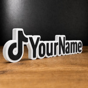 Peut inclure: Une enseigne en trois dimensions présentant l'icône noire du logo TikTok suivie du texte noir YourName, le tout sur une base blanche épaisse. Les lettres et l'icône ont une finition texturée et sont disposées horizontalement.
