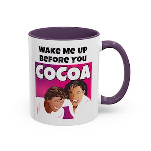 May include: White ceramic coffee mug with a purple rim and handle. The mug features a pink and purple background with a cartoon image of two men. The text on the mug reads "Wake Me Up Before You Cocoa".