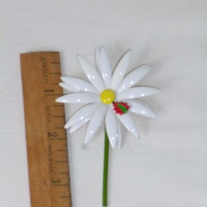 May include: White daisy brooch with a yellow center and a red and green holly leaf accent. The brooch is about 2 inches in diameter.