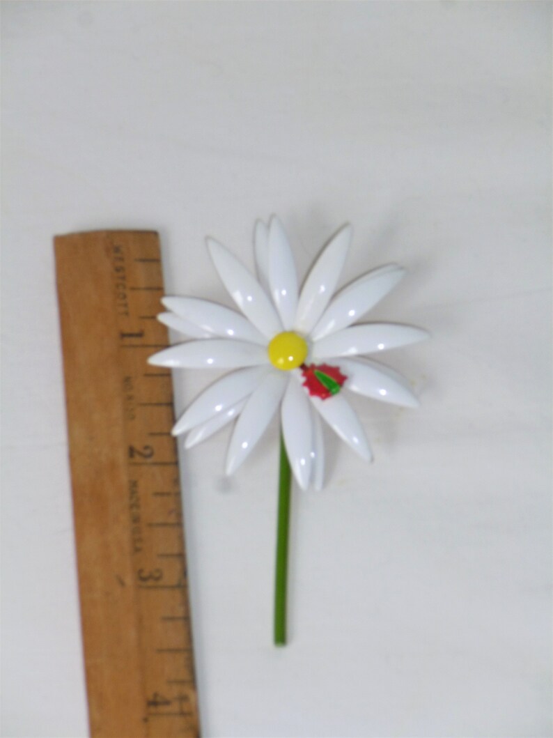 May include: White daisy brooch with a yellow center and a red and green holly leaf accent. The brooch is about 2 inches in diameter.
