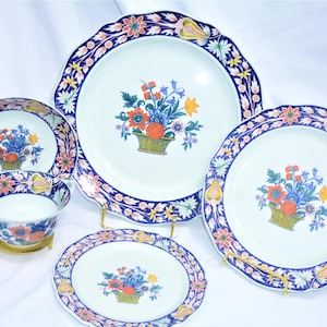 May include: A set of four white porcelain plates with blue and orange floral designs. The plates feature a scalloped edge and a basket of flowers in the center.