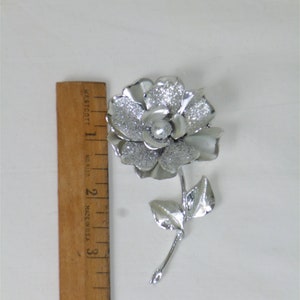 May include: A silver-toned brooch in the shape of a flower with a single pearl in the center. The flower has a textured surface and the stem has two leaves.