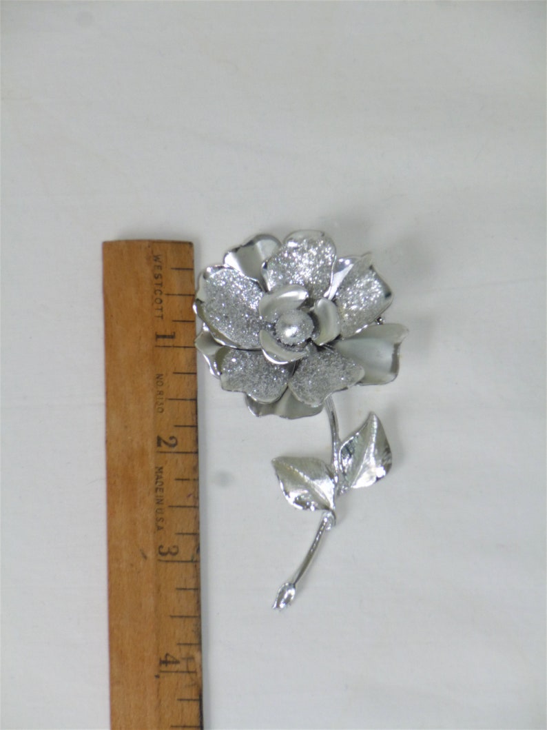 May include: A silver-toned brooch in the shape of a flower with a single pearl in the center. The flower has a textured surface and the stem has two leaves.