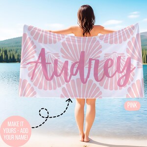 May include: A white beach towel with a pink seashell pattern and the name "Audrey" in pink script. The towel is held up by a person standing near a lake. The text "MAKE IT YOURS - ADD YOUR NAME!" is also visible.