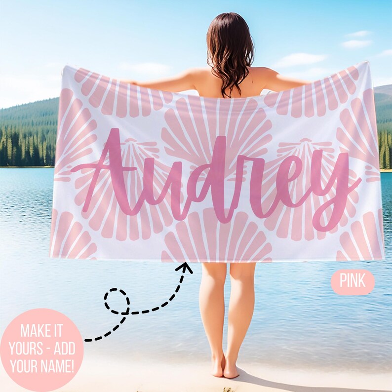 May include: A white beach towel with a pink seashell pattern and the name "Audrey" in pink script. The towel is held up by a person standing near a lake. The text "MAKE IT YOURS - ADD YOUR NAME!" is also visible.