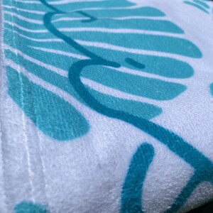May include: White beach towel with a teal leaf design. The towel has a soft, textured surface and a teal border. The leaf design is a repeating pattern of stylized leaves and vines.
