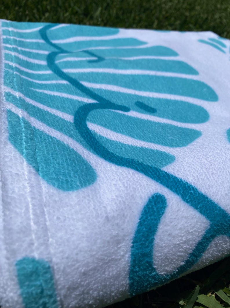 May include: White beach towel with a teal leaf design. The towel has a soft, textured surface and a teal border. The leaf design is a repeating pattern of stylized leaves and vines.