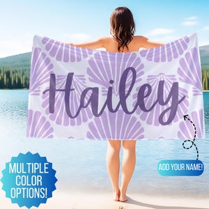 May include: A white beach towel with a repeating lavender seashell pattern and the name "Hailey" in a matching script font. The towel is held up by a person standing near a lake. Text on the image says "Multiple Color Options!" and "Add Your Name!"