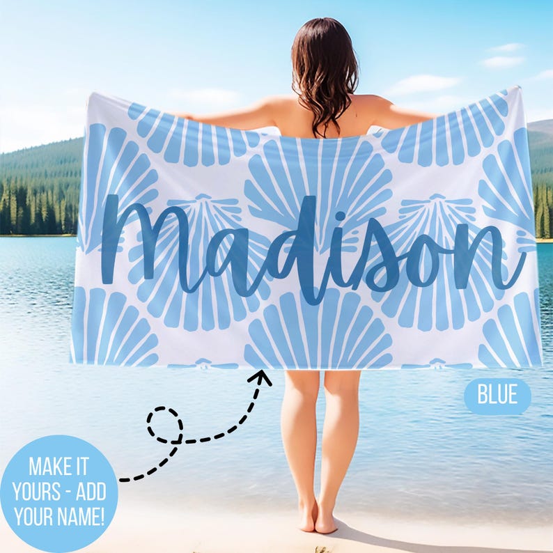 May include: A white and blue beach towel with a seashell pattern and the name "Madison" in blue script. The towel is held up in front of a lake. The word "BLUE" is in a blue box.