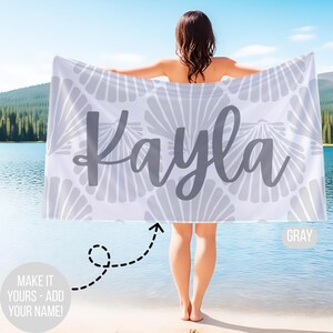May include: A white beach towel with a gray seashell pattern and the name "Kayla" in gray script. The towel is held up by a person standing near a lake. The text "MAKE IT YOURS - ADD YOUR NAME!" is also visible.