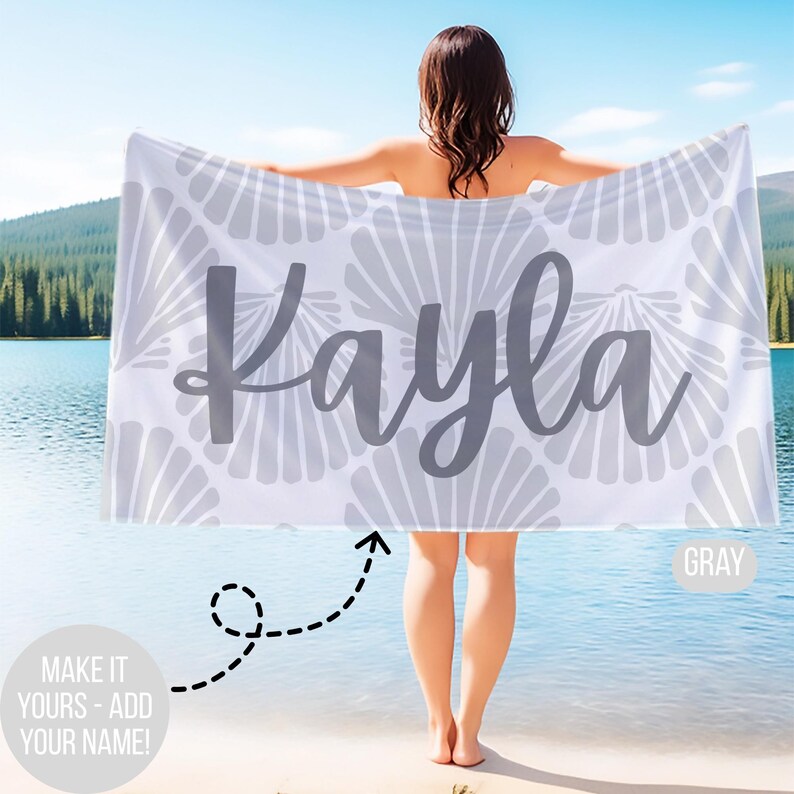 May include: A white beach towel with a gray seashell pattern and the name "Kayla" in gray script. The towel is held up by a person standing near a lake. The text "MAKE IT YOURS - ADD YOUR NAME!" is also visible.