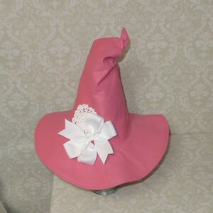 Puede incluir: Un sombrero de bruja rosa con un lazo blanco y un borde de encaje blanco.