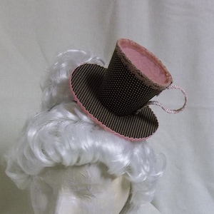 May include: A brown and white polka dot teacup hat with a pink trim and a pink interior. The hat is worn on a white wig.