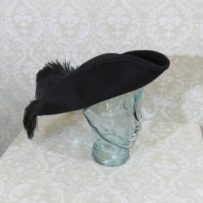 Puede incluir: Un sombrero tricornio negro con ala curva y penacho de plumas decorativo. El sombrero se exhibe sobre una cabeza de cristal transparente contra un fondo estampado. El ala del sombrero mide unos 30 cm.