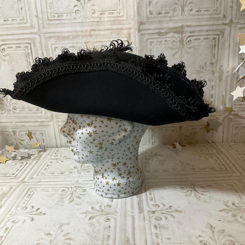 May include: A black tricorn hat with a wide brim and decorative trim, displayed on a mannequin head. The hat features a curved brim and is adorned with a delicate trim along the edges. The mannequin head is decorated with gold stars.