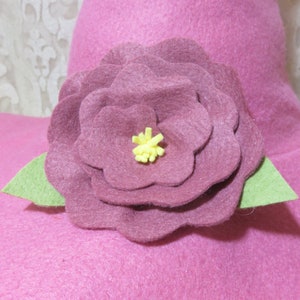 Puede incluir: Una flor de fieltro rosa con un centro amarillo, acentuada con hojas de fieltro verde. La flor est&aacute; unida a una superficie de fieltro rosa.