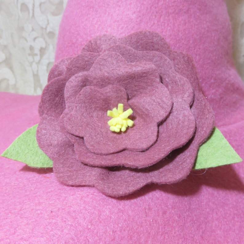 Puede incluir: Una flor de fieltro rosa con un centro amarillo, acentuada con hojas de fieltro verde. La flor est&aacute; unida a una superficie de fieltro rosa.