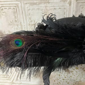 May include: Black hat adorned with peacock feathers and black plumes. The hat features a prominent peacock feather with a vibrant blue and green eye. The hat's design includes a mix of textures and shapes, creating a dramatic and elegant look.