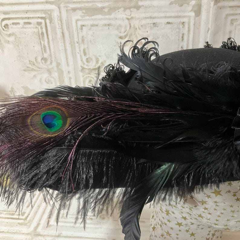 May include: Black hat adorned with peacock feathers and black plumes. The hat features a prominent peacock feather with a vibrant blue and green eye. The hat's design includes a mix of textures and shapes, creating a dramatic and elegant look.