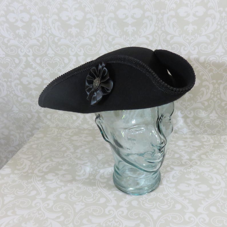 Puede incluir: Un sombrero tricornio negro con una flor decorativa negra y un centro plateado. El sombrero se exhibe sobre una forma de cabeza de vidrio transparente. El sombrero tiene un ala curva y un ribete decorativo.