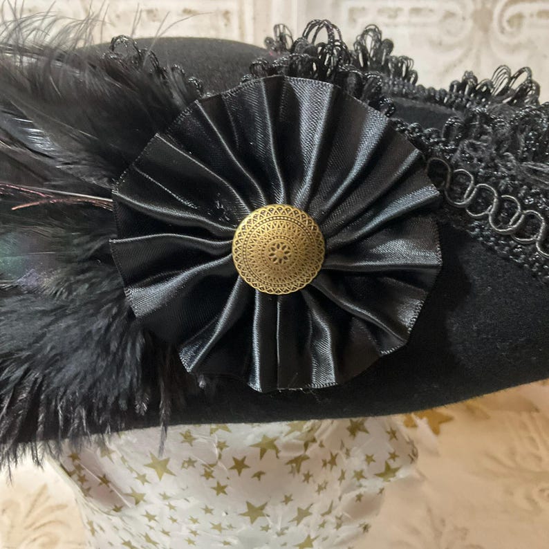 May include: A black hat with a decorative black satin rosette and a gold-colored button in the center. The hat is adorned with black feathers and lace trim, creating a vintage aesthetic.
