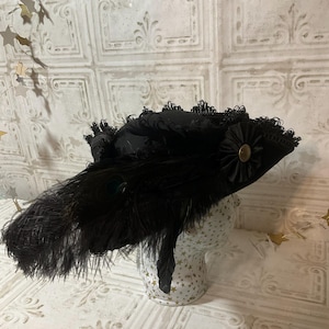 May include: A black hat with a wide brim, adorned with feathers and lace. The hat features a decorative flower with a gold-colored center and is displayed on a mannequin head. The hat's design is reminiscent of vintage fashion.