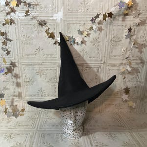 May include: A black witch hat with a wide brim and a tall, pointed crown. The hat is displayed on a decorative head form with a star pattern. The background features a garland of star-shaped decorations.