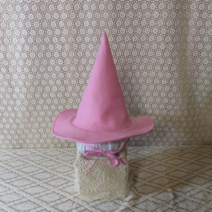 K&ouml;nnte beinhalten: Ein rosa Hexenhut mit breiter Krempe und hoher, spitzer Krone. Der Hut ist aus Filz gefertigt und sitzt auf einer wei&szlig;en Stoffbasis, die mit einem rosa Band gebunden ist. Der Hintergrund zeigt einen cremefarbenen Stoff mit Muster.