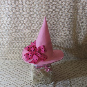 K&ouml;nnte beinhalten: Ein rosa Hexenhut mit breiter Krempe und spitzer Spitze. Der Hut ist mit einer gro&szlig;en rosa Schleife mit schwarzen Punkten verziert. Eine kleinere rosa Schleife ist um die Basis des Hutes gebunden.