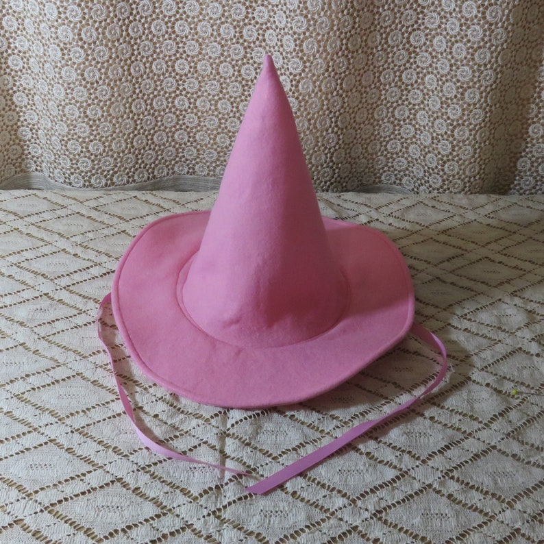 K&ouml;nnte beinhalten: Ein rosa Hexenhut mit breiter Krempe und spitzer Krone. Der Hut ist aus einem weichen Material gefertigt und hat ein passendes rosa Band als Kinnriemen. Der Hut ist f&uuml;r Kost&uuml;m- oder Anziehspiele gedacht.