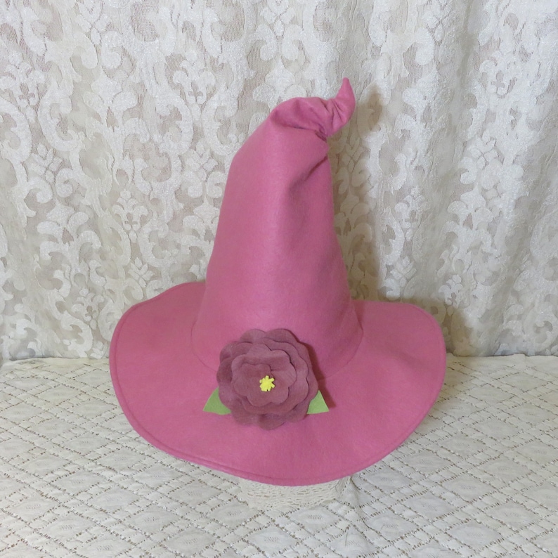 Puede incluir: Un sombrero de bruja de fieltro rosa con una gran flor de fieltro rosa y hojas de fieltro verde.