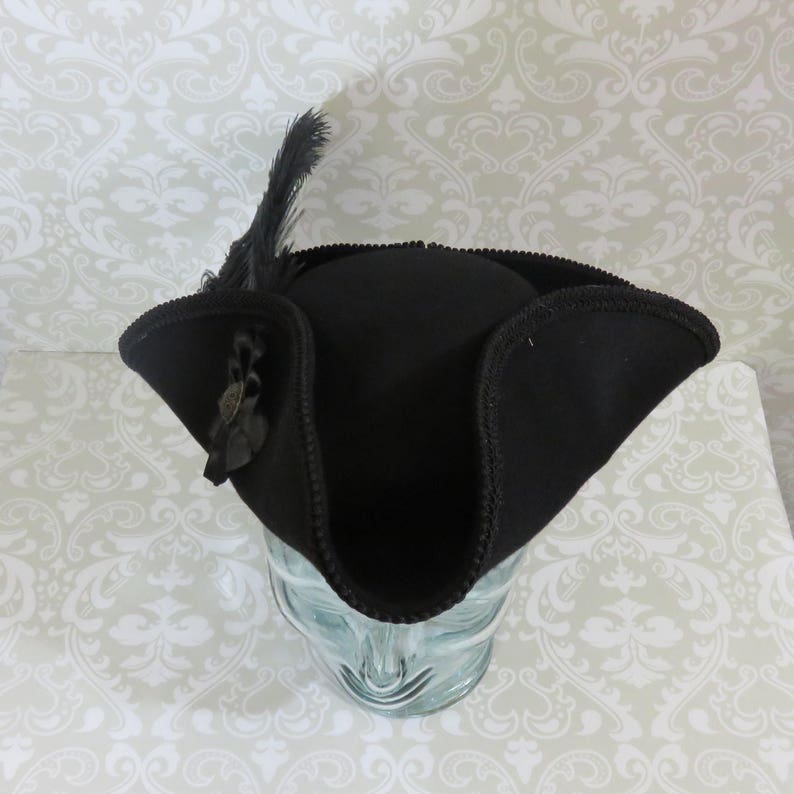 Puede incluir: Un sombrero tricornio negro con ala curva y una pluma decorativa. El sombrero tiene un borde cosido y un peque&ntilde;o adorno en el lateral. El sombrero se exhibe sobre una cabeza de cristal transparente.