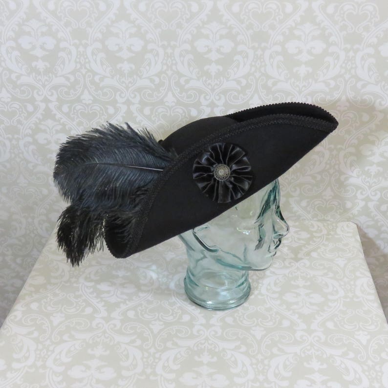 Puede incluir: Un sombrero tricornio negro con una roseta decorativa de sat&eacute;n negro y un bot&oacute;n central de estilo antiguo. El sombrero presenta un gran penacho de plumas negras y se exhibe sobre una cabeza de cristal transparente.