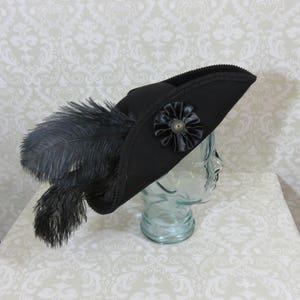 Puede incluir: Un sombrero tricornio negro con una flor decorativa de sat&eacute;n negro y un bot&oacute;n de bronce. El sombrero est&aacute; adornado con plumas negras y un ribete decorativo. El sombrero se exhibe sobre una cabeza de cristal.