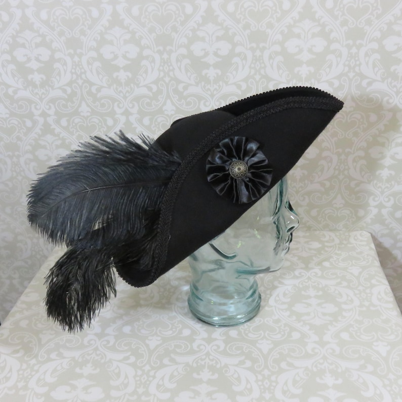 Puede incluir: Un sombrero tricornio negro con una flor decorativa de sat&eacute;n negro y un bot&oacute;n de bronce. El sombrero est&aacute; adornado con plumas negras y un ribete decorativo. El sombrero se exhibe sobre una cabeza de cristal.